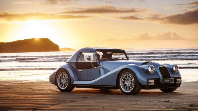 Blue vintage-style sports car parked on a sandy beach at sunset with ocean waves in the background.