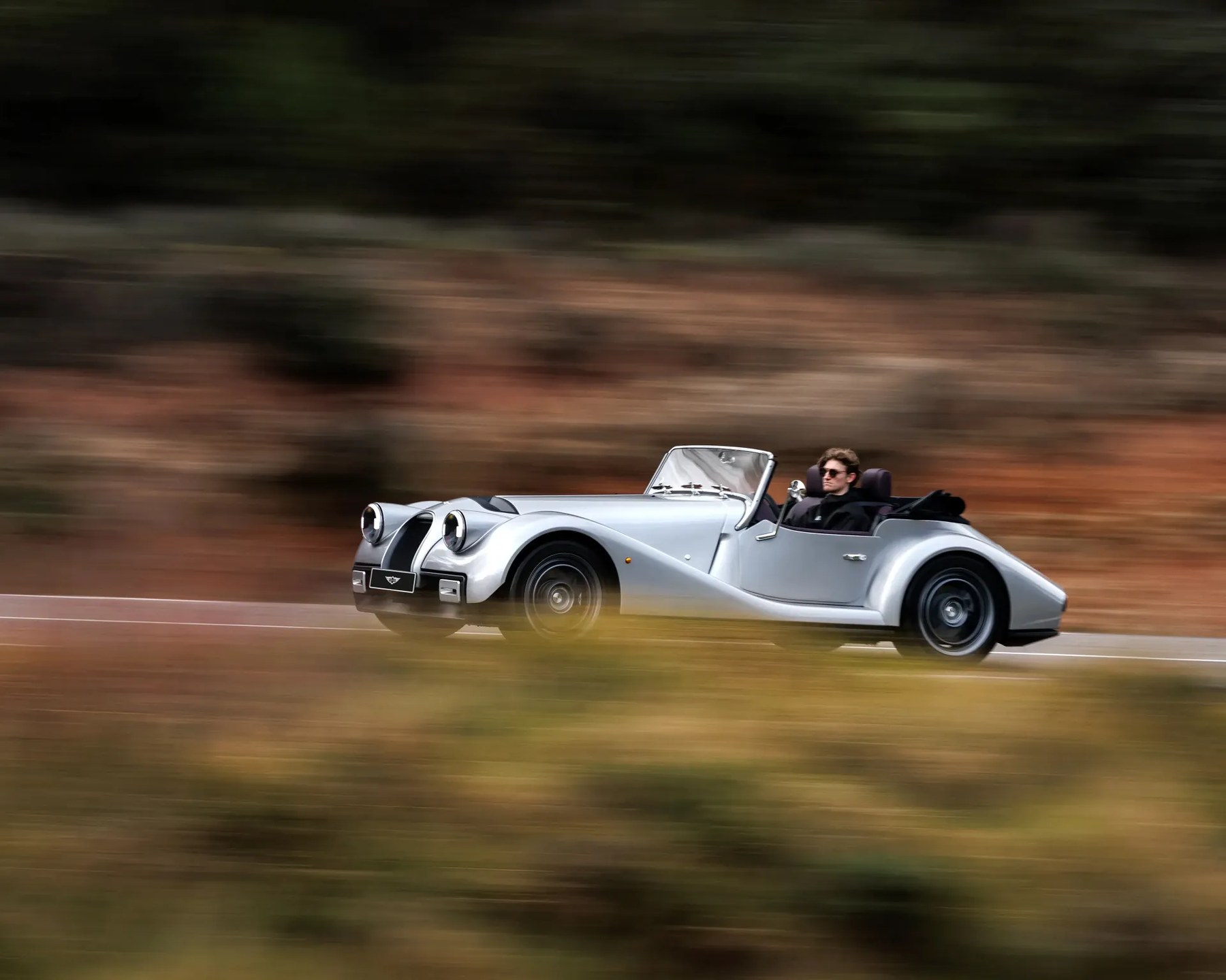 Silver classic convertible car driving on a road with blurred natural background.