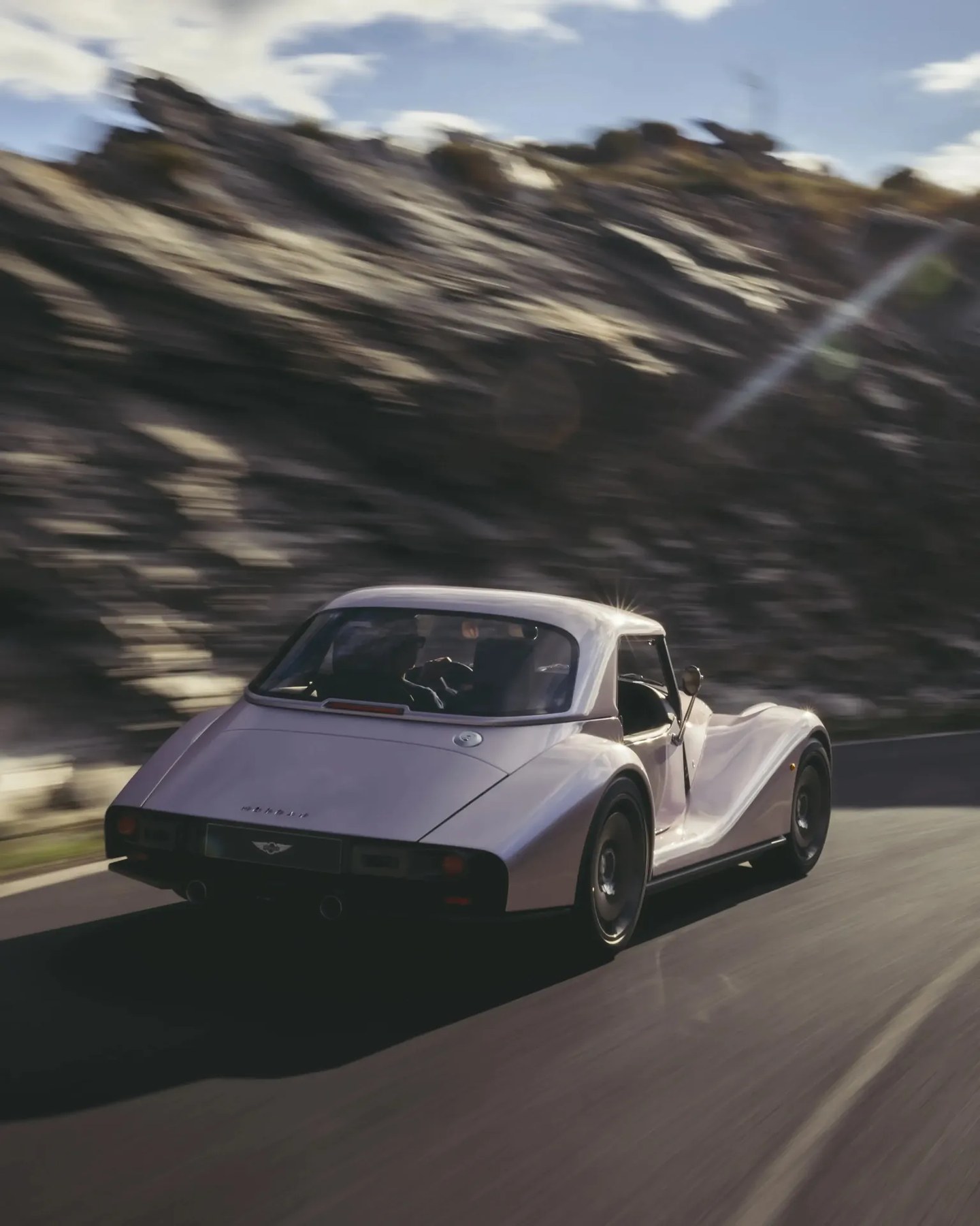 White Morgan 4/4 classic sports car driving on a road with rocky hillside in the background.